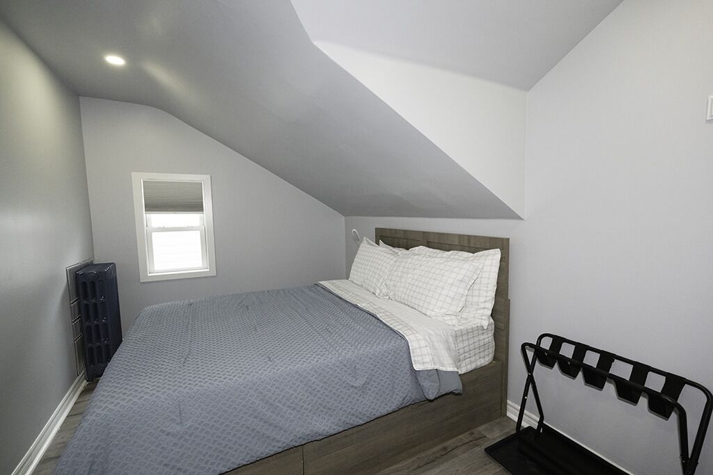 A bedroom with a sloped ceiling, a bed with white bedding, a window, and a radiator.