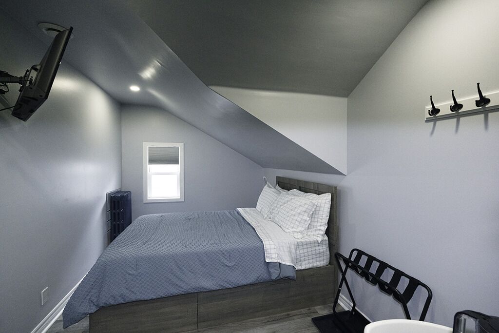 A bedroom with a sloped ceiling, a bed with a gray and white checkered bedspread, a black metal headboard, a small window, and a wall-mounted shelf with three black objects.