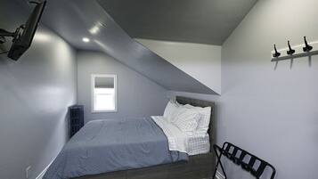 A bedroom with a sloped ceiling, a bed with a gray and white checkered bedspread, a black metal headboard, a small window, and a wall-mounted shelf with three black objects.