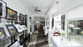 A spacious hallway with white walls, a wooden floor, and a ceiling fan. There are framed pictures on the left and a table with a vase of flowers on the right.