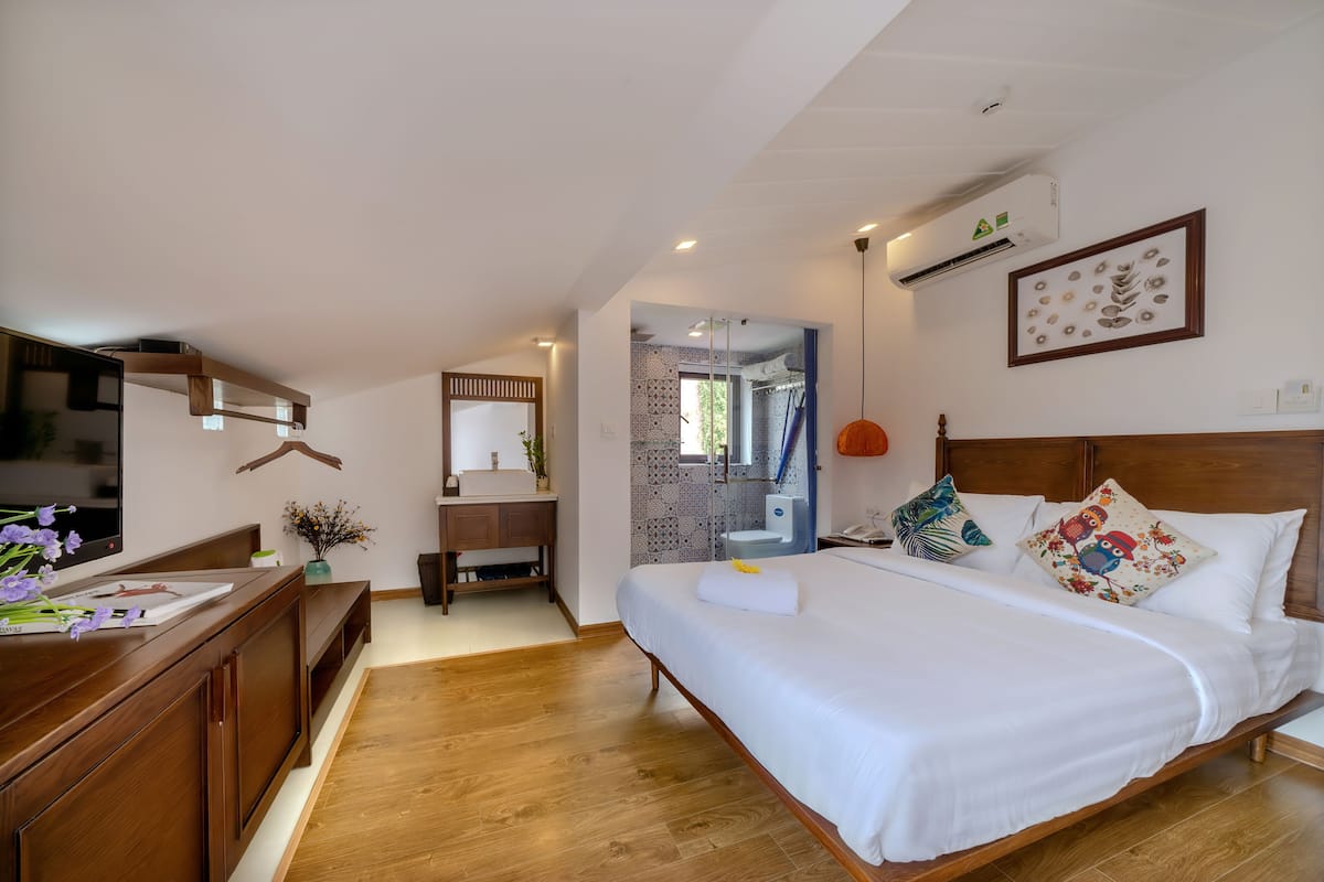 Een slaapkamer met een houten bed, wit beddengoed, een bureau met een computer, en een badkamer die door een open deur zichtbaar is.