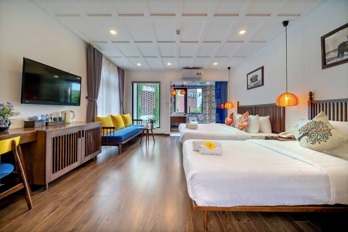 Hotelkamer met een groot flatscreen televisietoestel, een bank, twee bedden met kussens, een houten vloer en een plafond met inbouwverlichting.