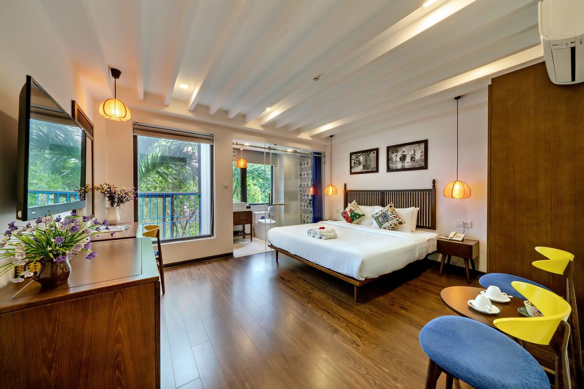 Een moderne hotelkamer met een groot bed, houten vloeren, een bureau met een bloevas en uitzicht op het omliggende landschap.