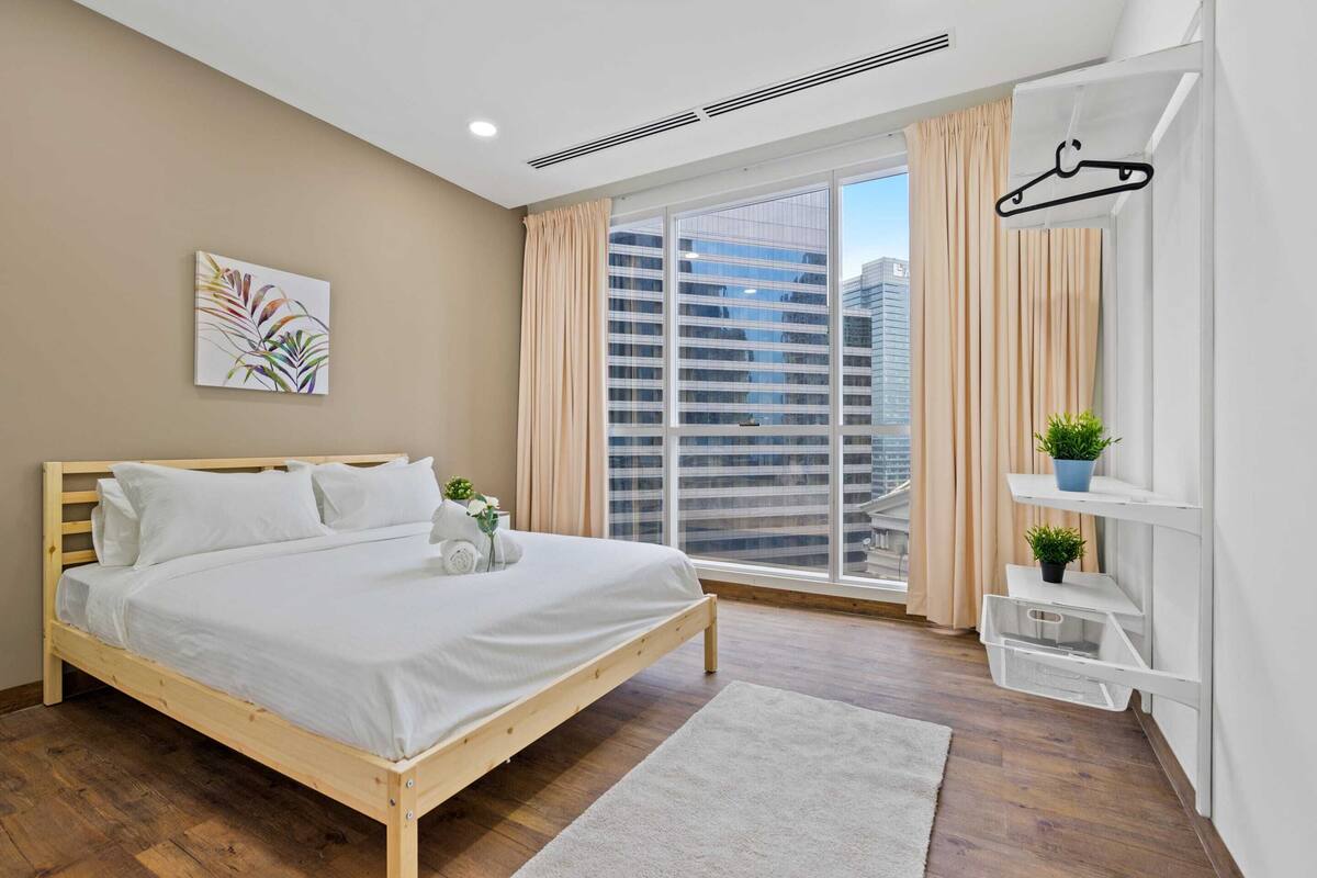 A modern bedroom with a wooden bed, a large window with curtains, a view of buildings, and a small shelf with plants.