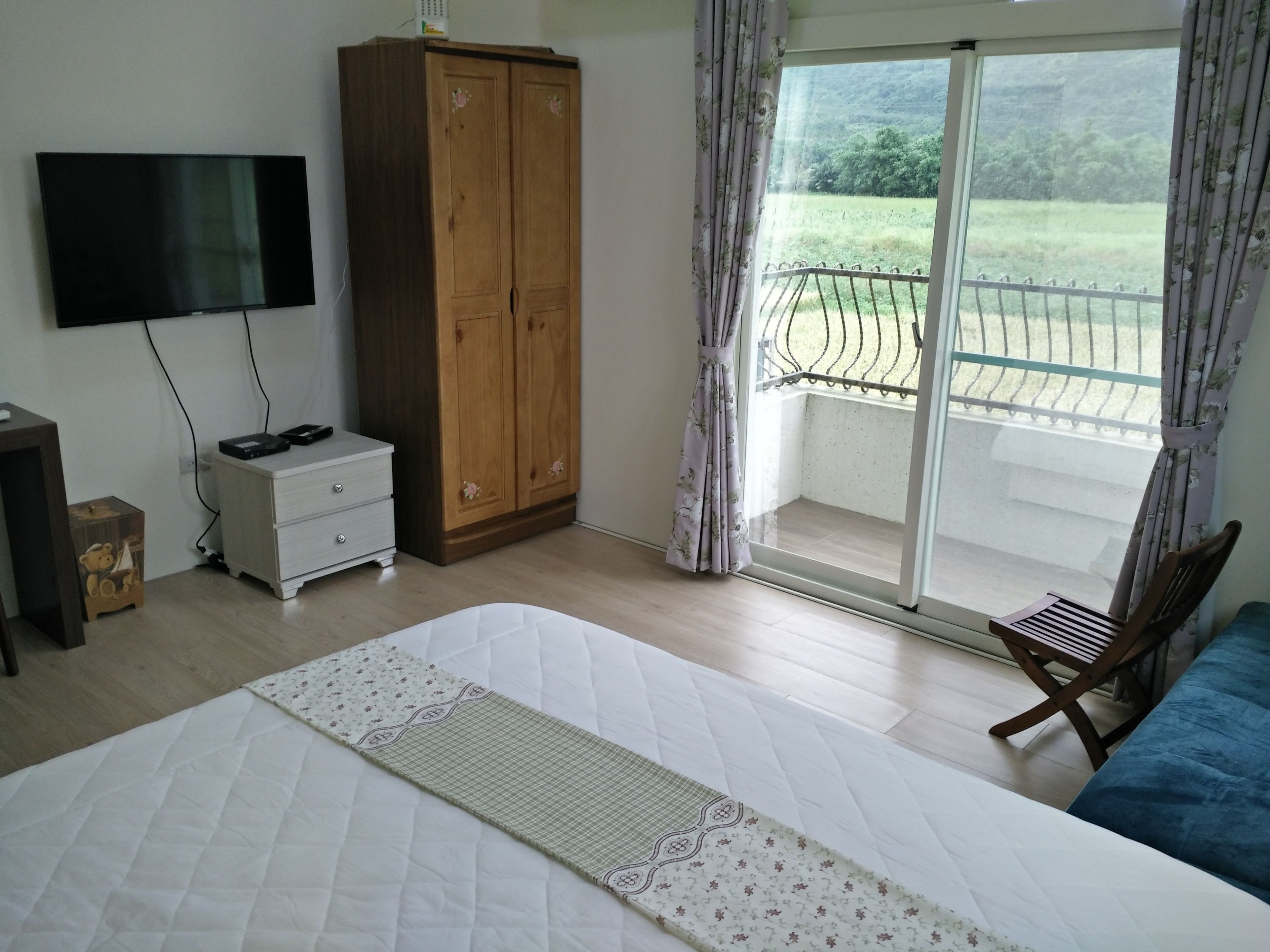A bedroom with a large bed, a wardrobe, a television, and a balcony with a view of greenery.