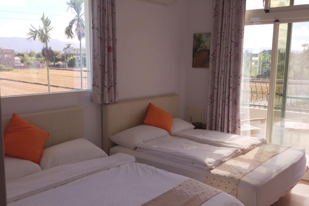 Habitación de hotel con tres camas individuales, cada una con ropa de cama blanca y almohadas naranjas. La habitación tiene un ventanal con cortinas florales, un cuadro en la pared y vistas a palmeras y edificios.