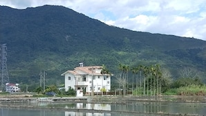 Una casa de dos pisos con techo de tejas, rodeada por una masa de agua y palmeras, con una montaña al fondo.