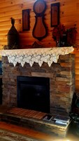 Uma lareira de pedra com um mantel de renda, um relógio e quadros nas paredes acima.