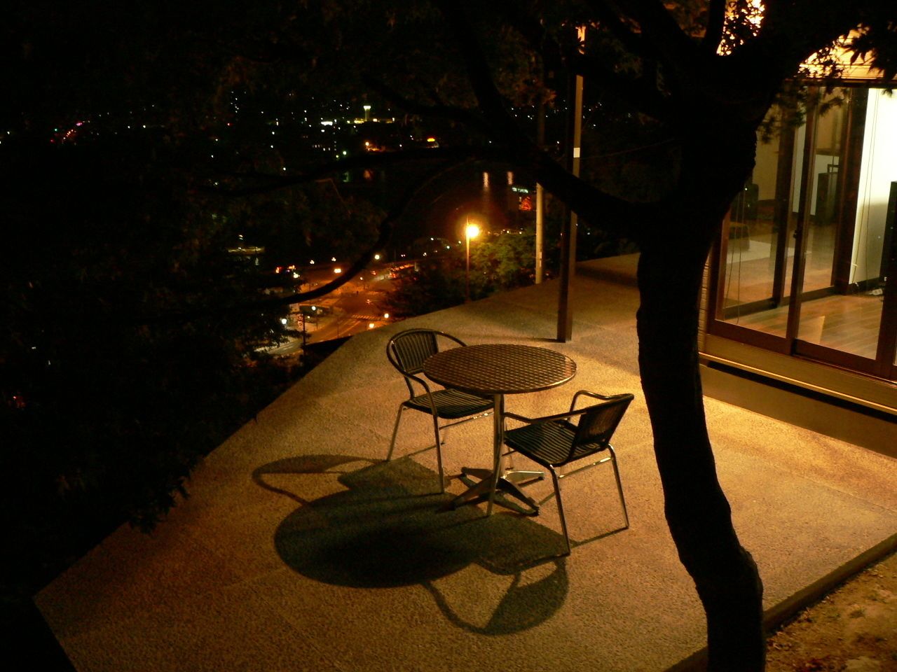 A nighttime outdoor seating area with a table and chairs, a tree, and a view of a cityscape with lights.