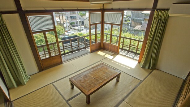 Une pièce avec un sol recouvert de tatamis, une table en bois, de grandes fenêtres et une vue sur un quartier résidentiel.