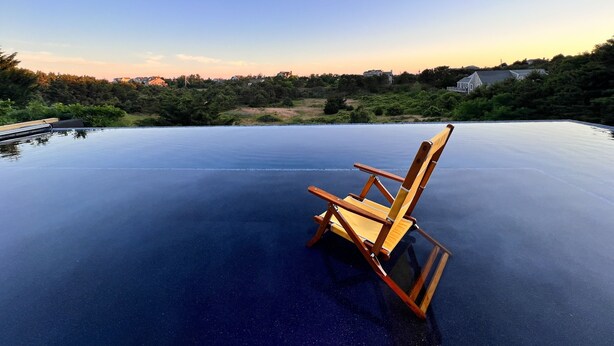 Uma cadeira de madeira em uma piscina infinita de vidro, com vista para casas e árvores ao fundo.