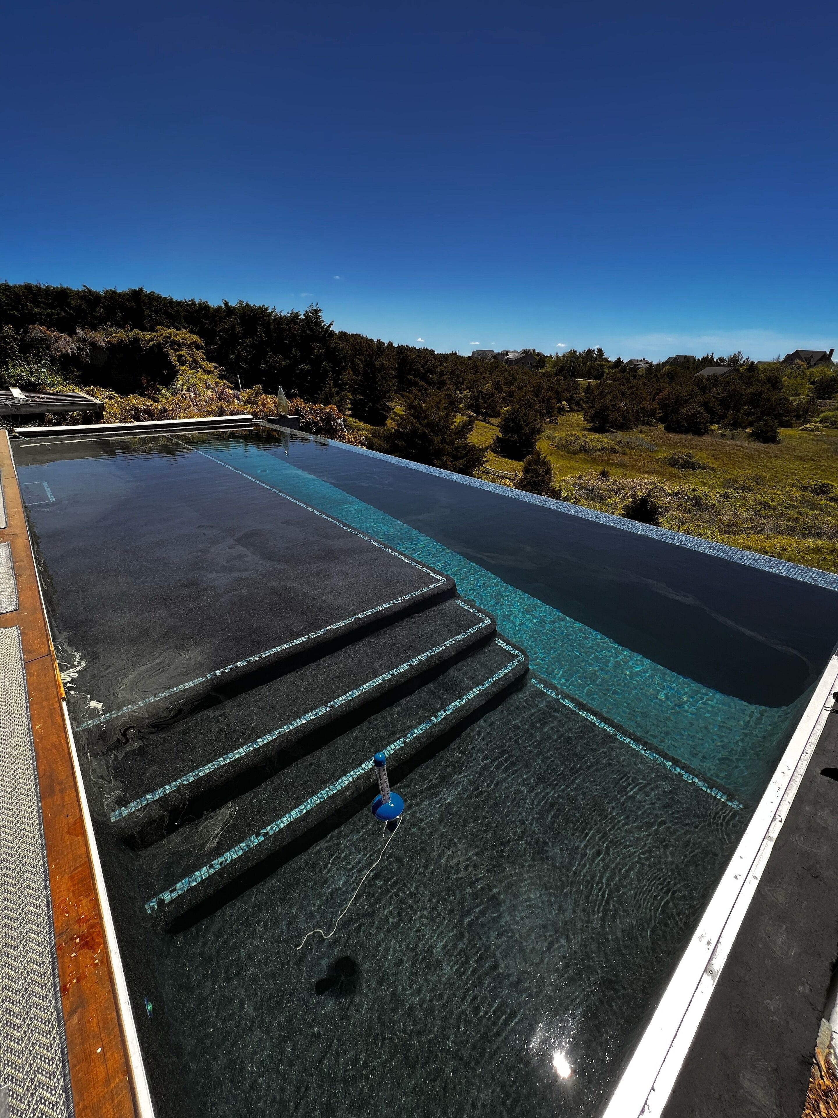 A rooftop infinity pool with clear water, steps leading to the edge, and a view of a forested landscape.