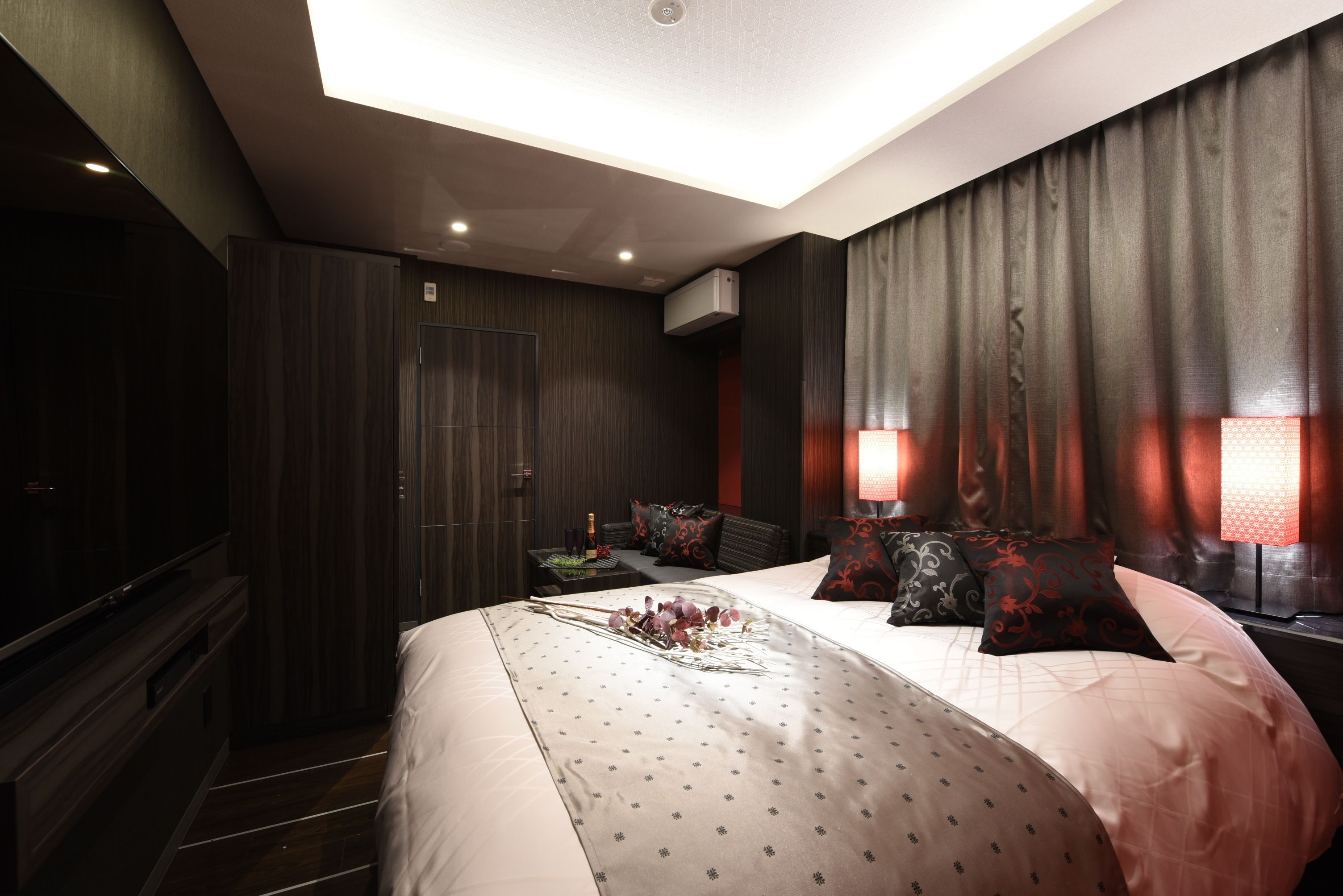 A modern hotel room with a large bed, dark wood paneling, a flat-screen TV, and a ceiling light.