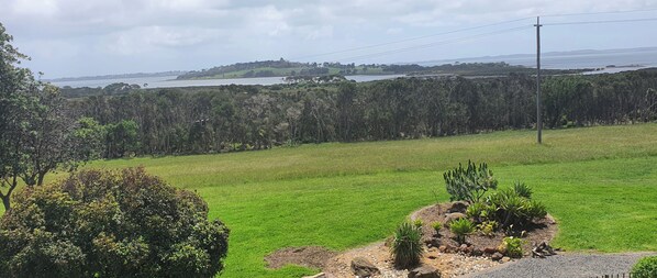 Un campo cubierto de hierba con una pequeña zona de jardín, un bosque frondoso al fondo y una masa de agua en el horizonte.