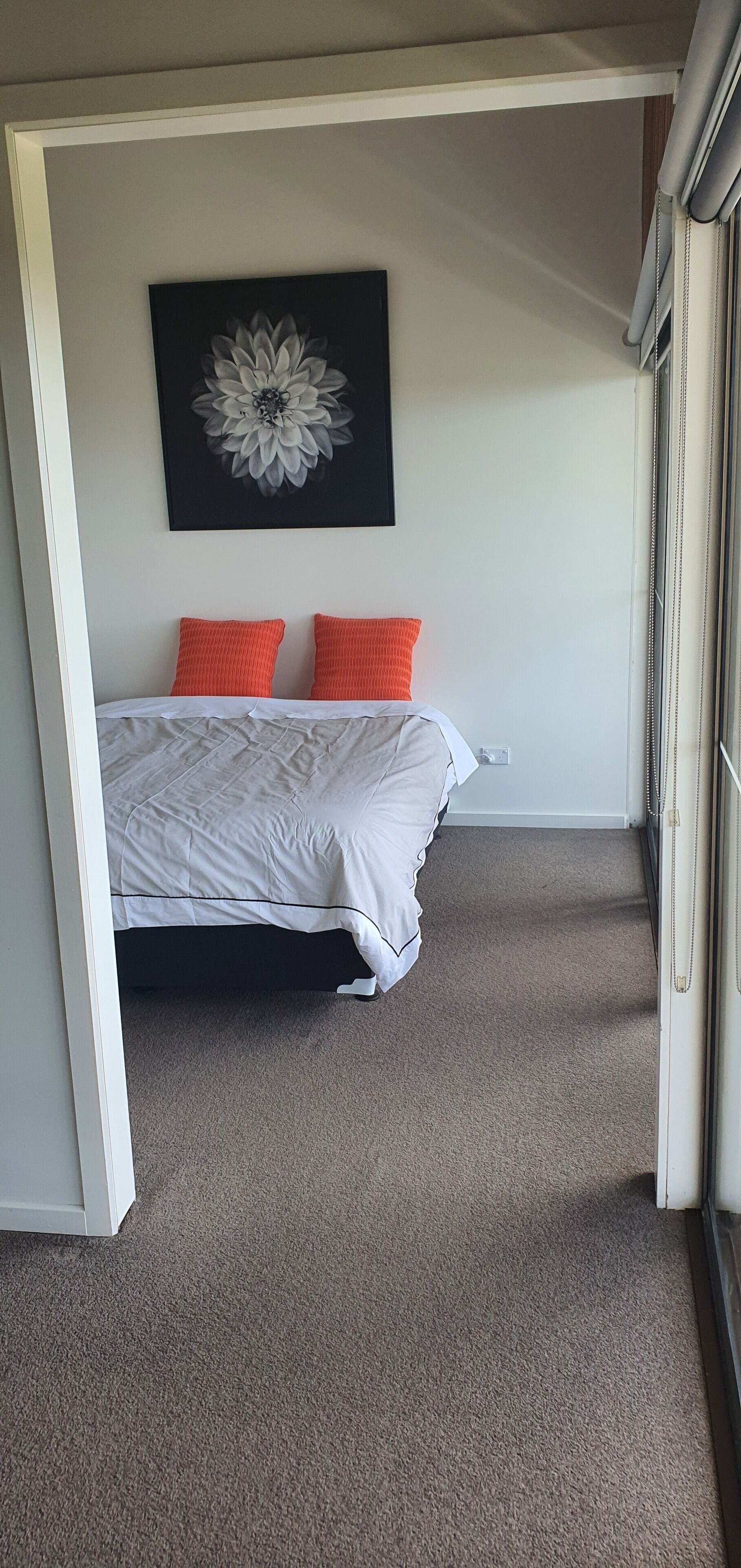 Un dormitorio con una cama, dos almohadas naranjas, una colcha blanca y una obra de arte floral blanco y negro en la pared.