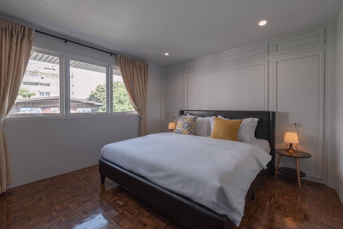 Un dormitorio ordenado con una cama grande, dos lámparas de noche y vistas a edificios a través de una ventana con cortinas.