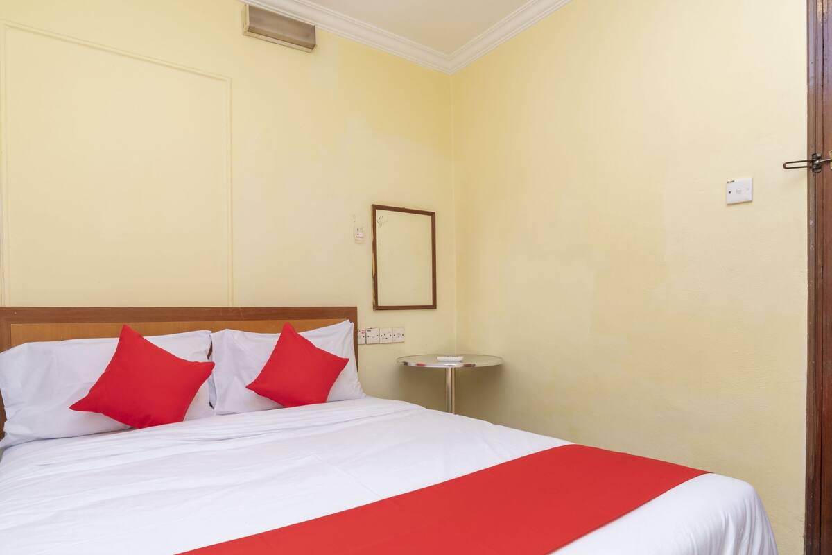 Une chambre d’hôtel avec un lit, une literie rouge et blanche, une petite table de chevet et un miroir fixé au mur.