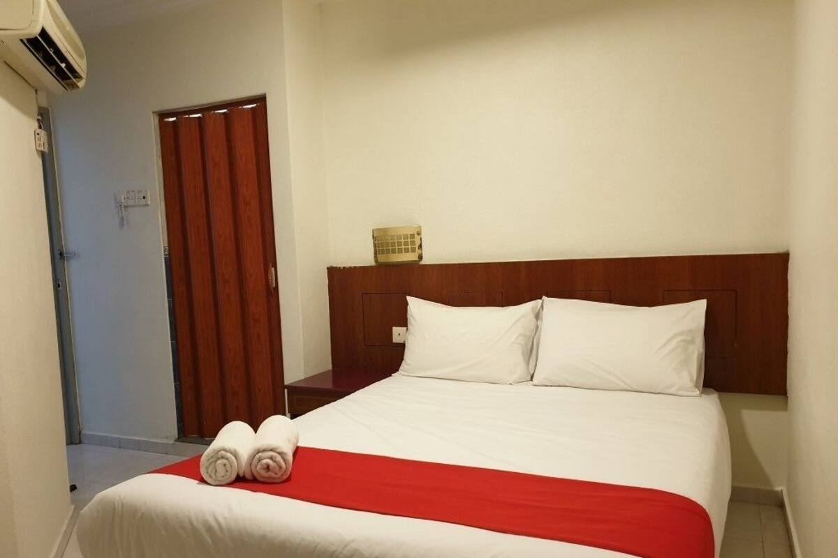 Une chambre d’hôtel avec un lit, une couvre-lit rouge et blanche, deux serviettes enroulées, une tête de lit en bois et une petite table de chevet avec une lampe.
