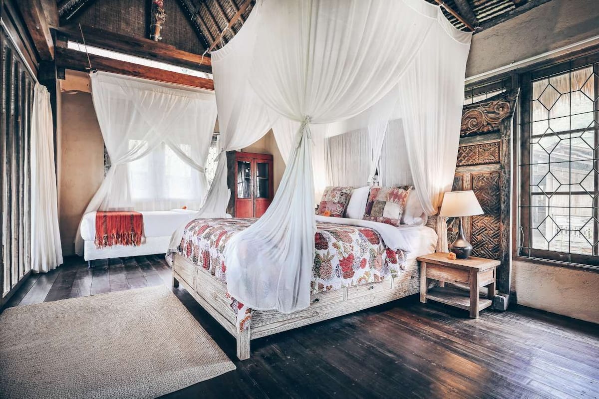 A bedroom with a four-poster bed, a nightstand, a window with a geometric pattern, and a wooden ceiling.