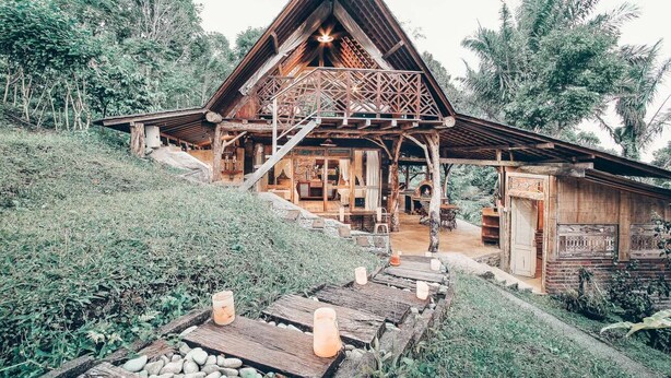 Une cabane en bois avec un toit triangulaire, une véranda dotée de balustrades en bois et un chemin de pierre menant à l’entrée.