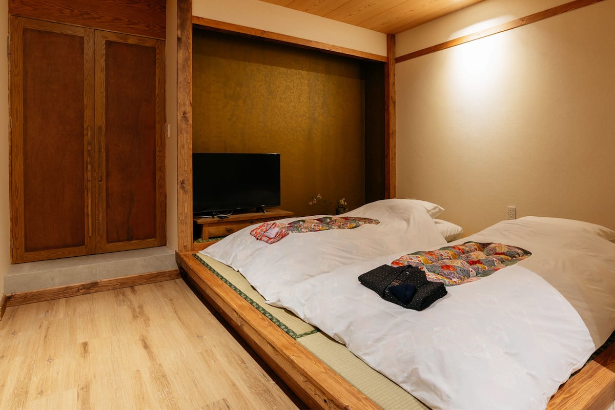 A traditional Japanese-style room with a tatami mat floor, a wooden wardrobe, a television, and a bed with pillows and a quilt.