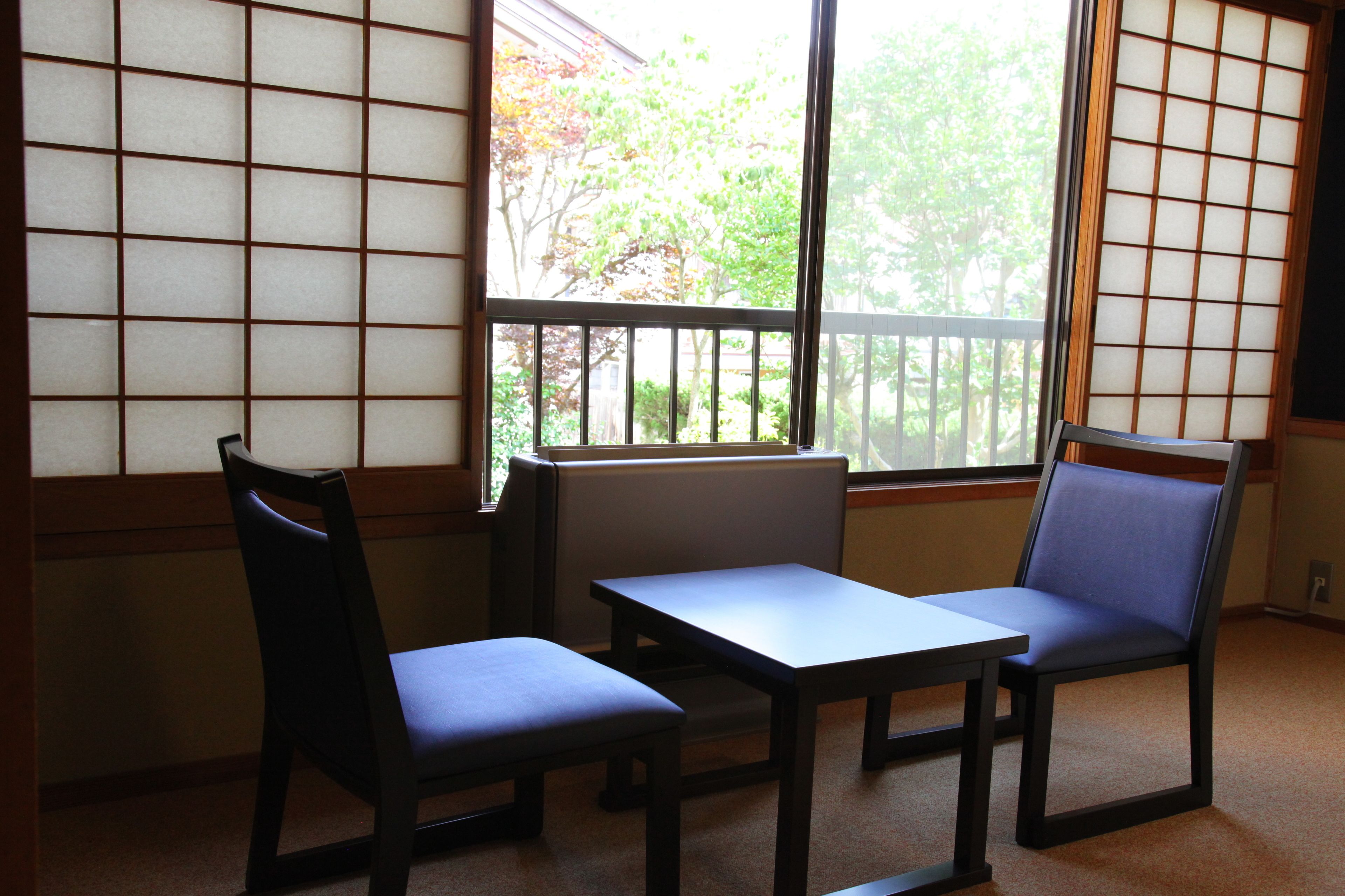 Una habitación con suelo de esteras de tatami, una mesita baja, dos sillas y un gran ventanal con vistas a árboles.