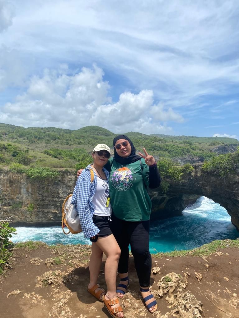 Two people standing on a rocky outcrop with a view of a coastal area and greenery in the background.