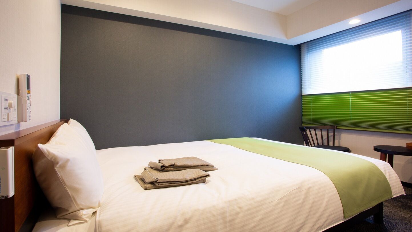 A hotel room with a bed, a desk, a chair, a window with blinds, and a wall-mounted control panel.