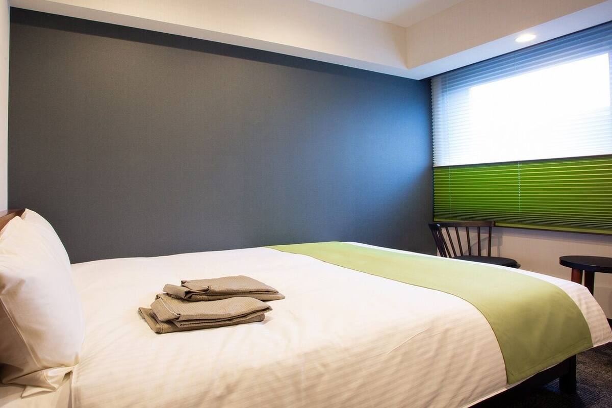A hotel room with a bed, a desk, a chair, a window with blinds, and a wall-mounted control panel.