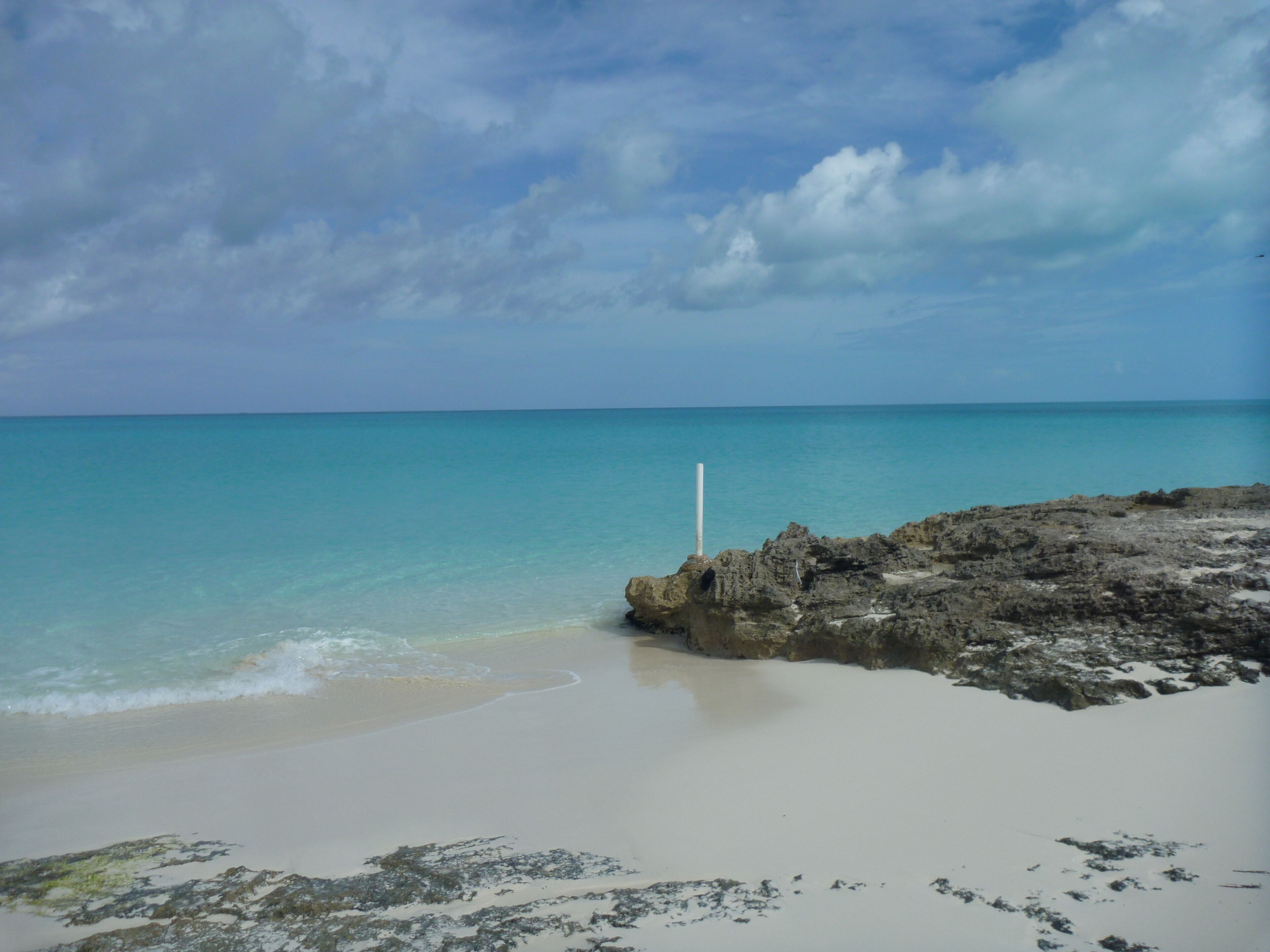 A beach with clear turquoise water, a rocky outcrop, and a white pole.