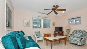 A living room with a white sofa, teal cushions, a wooden coffee table, a white armchair, a floral armchair, a TV, and a ceiling fan. - Ixora West - Views over Elizabeth harbor, beach access across the road. (George Town)