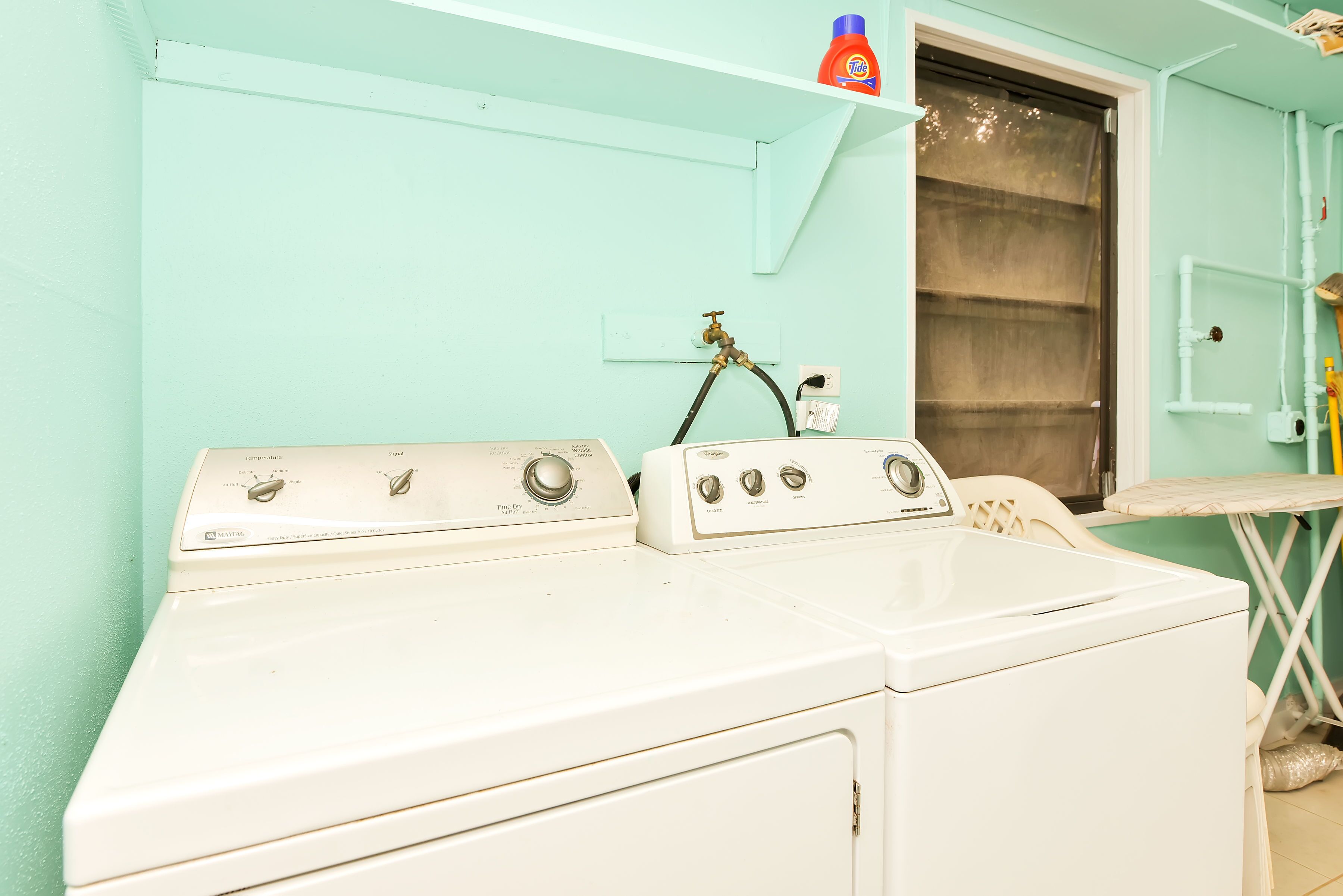 A laundry area with a white washer and dryer, a window with a curtain, and a shelf with a bottle of Tide detergent.