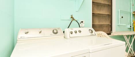 A laundry area with a white washer and dryer, a window with a curtain, and a shelf with a bottle of Tide detergent.