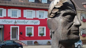 Ein Gebäude mit roter Fassade und weißen Fensterläden, davor ein schwarzes Auto und eine Steinbüste.