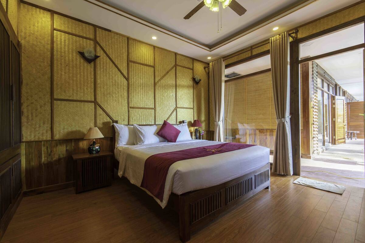 A hotel room with a wooden floor, a large bed with a headboard, a nightstand with a lamp, and a view of the outside through a glass door.
