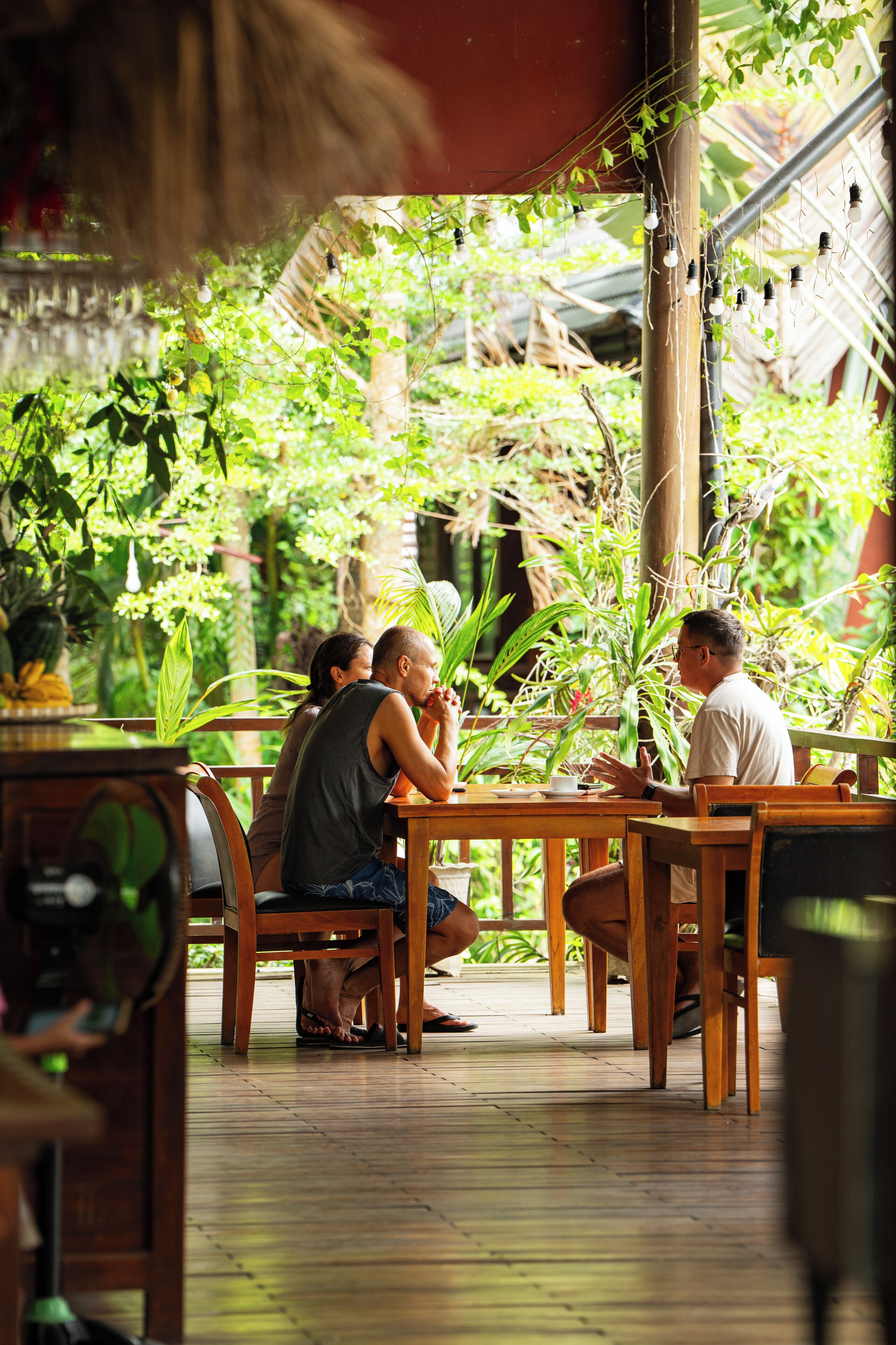 A wooden outdoor dining area with tables and chairs, surrounded by lush greenery and plants.