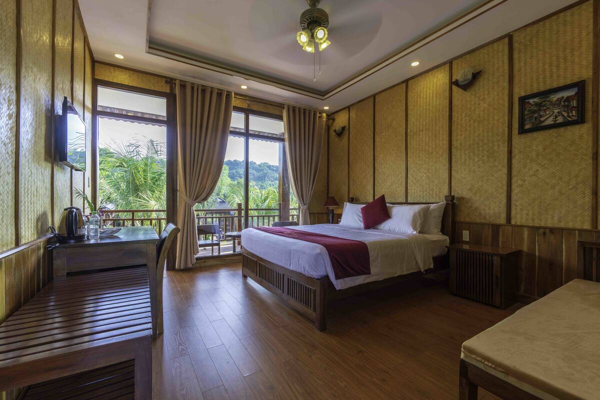 A hotel room with a bed, a desk, and a view of greenery outside.