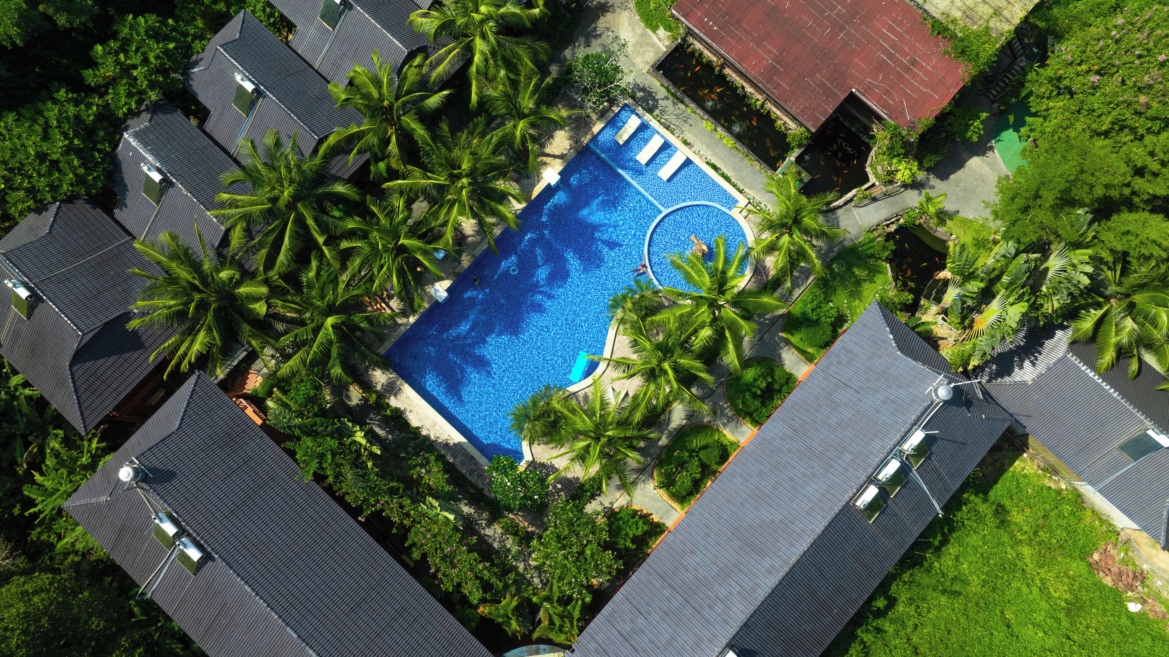 An aerial view of a resort with a central swimming pool surrounded by palm trees and multiple buildings with dark roofs.