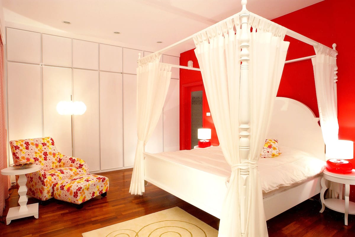 A bedroom with a four-poster bed, a floral armchair, and a bedside table with a lamp.