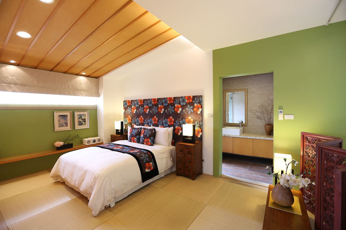 A bedroom with a bed, bedside tables, a nightstand, a mirror, and a bathroom visible through an open door.
