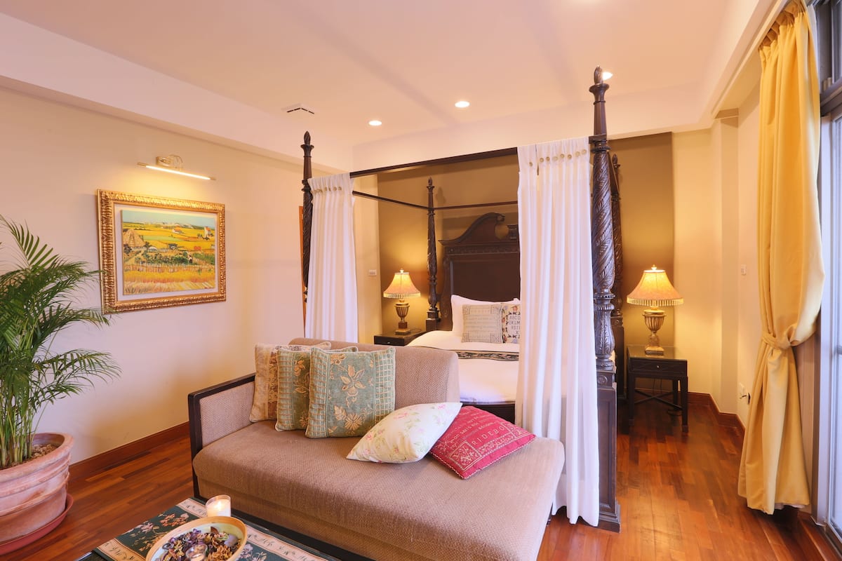 A four-poster bed with a canopy, a sofa with decorative pillows, a potted plant, and a framed painting on the wall.