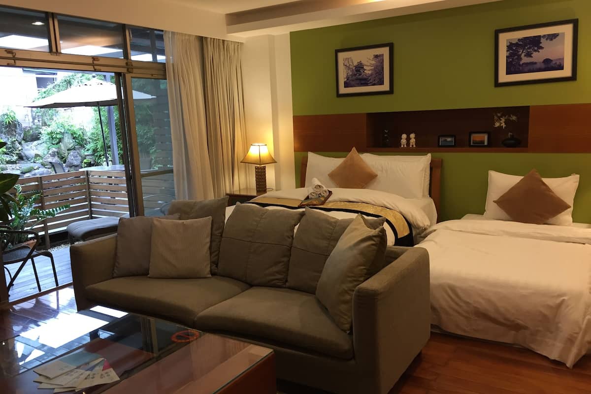 A hotel room with a sofa, a bed, a glass coffee table, and a view of a garden through a sliding door.