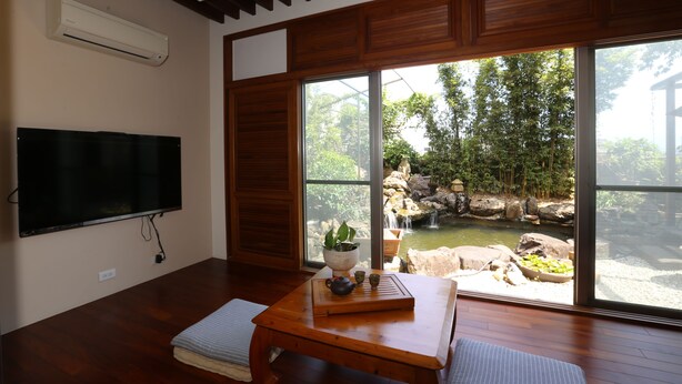 A room with a flat-screen TV, a wooden table with a tray, a potted plant, and a view of a pond and trees outside. - Memory Garden Villa (Luodong)