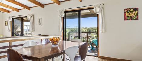 A dining area with a wooden table and chairs, a bowl of fruit, and a view of the outdoors through large windows.