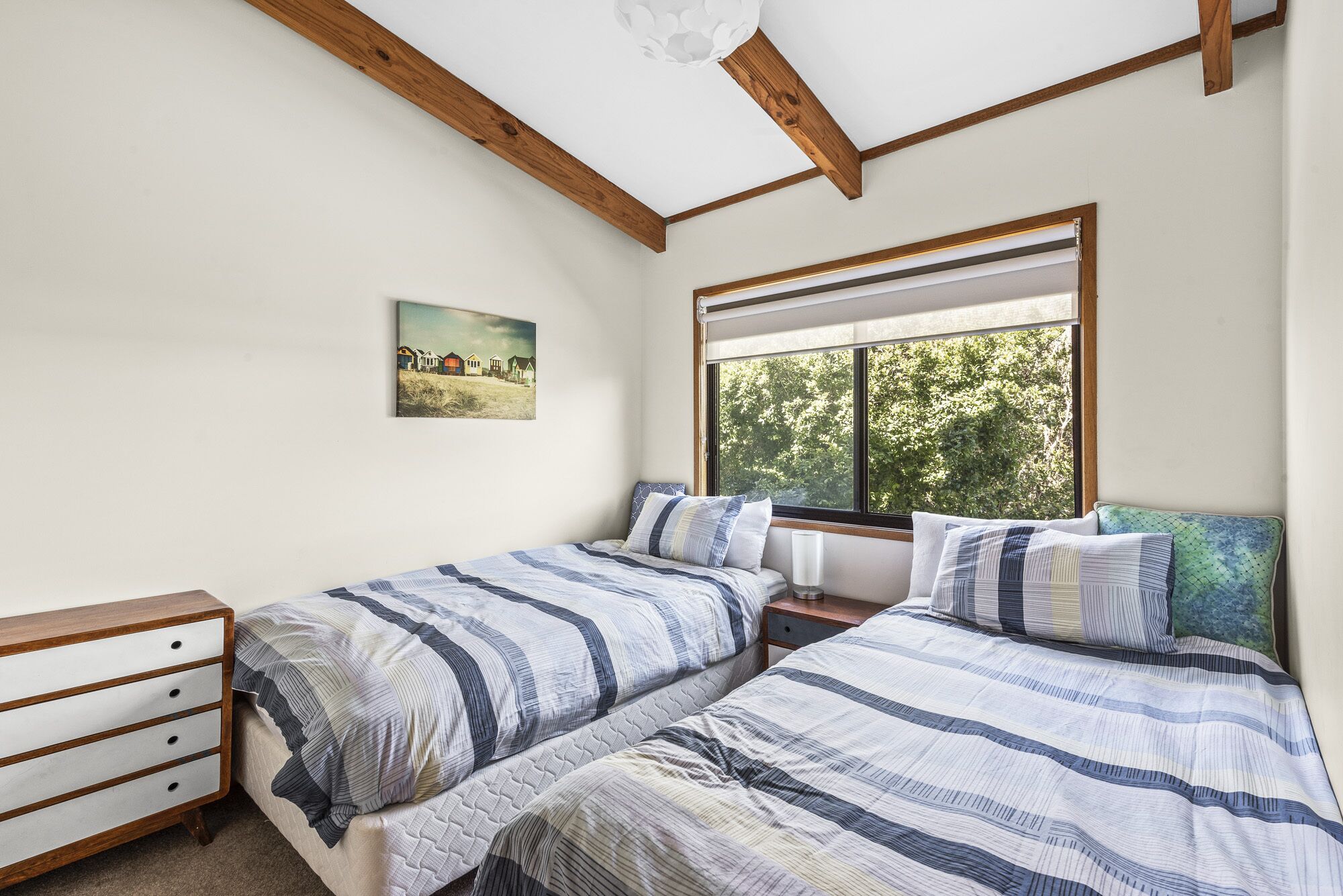 A bedroom with two beds, a wooden dresser, a painting, and a window with a view of trees.