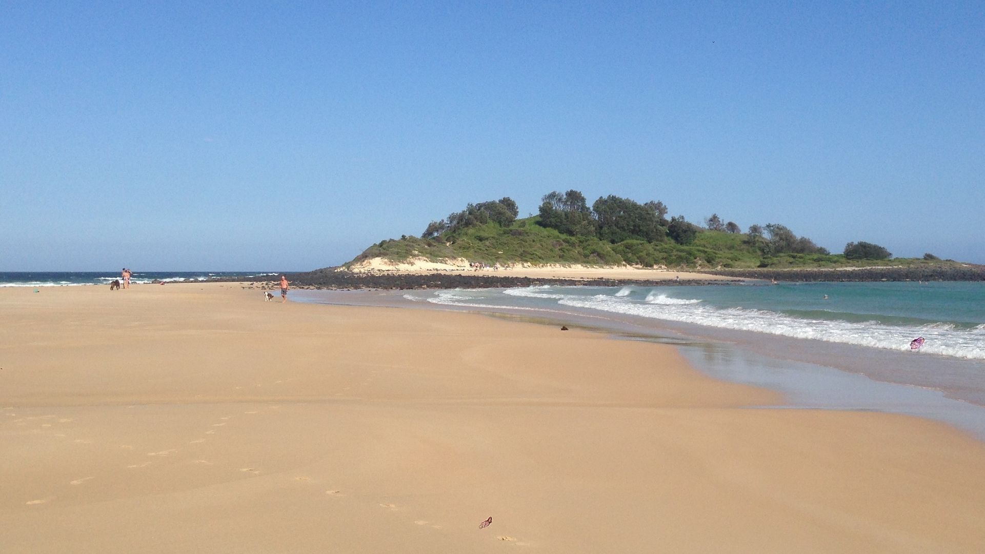 A wide sandy beach with gentle waves, a small hill with trees, and a clear sky.