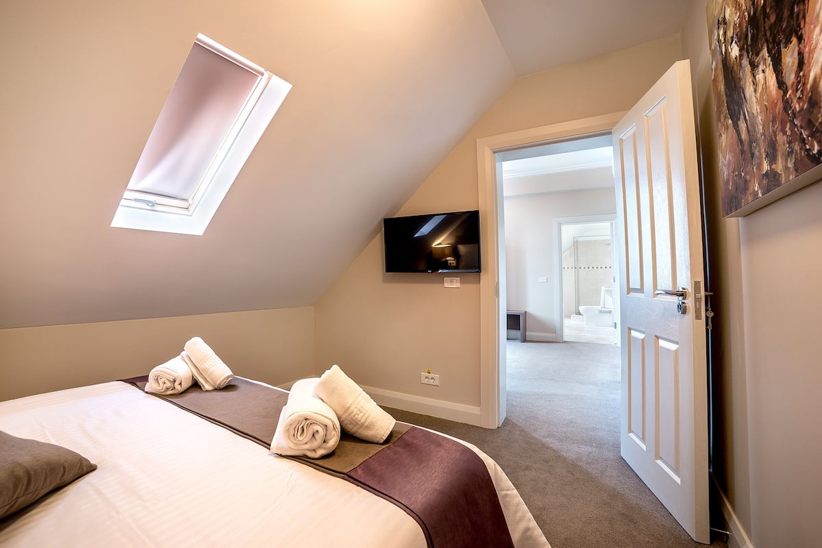 Quarto de hotel com uma cama, duas toalhas enroladas, uma televisão na parede e uma porta que dá para outro quarto.