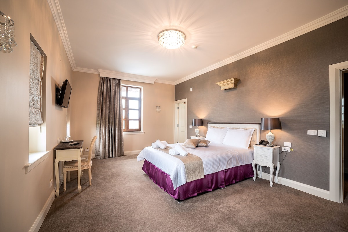 Quarto com cama, mesa de cabeceira, abajur e janela com cortinas.