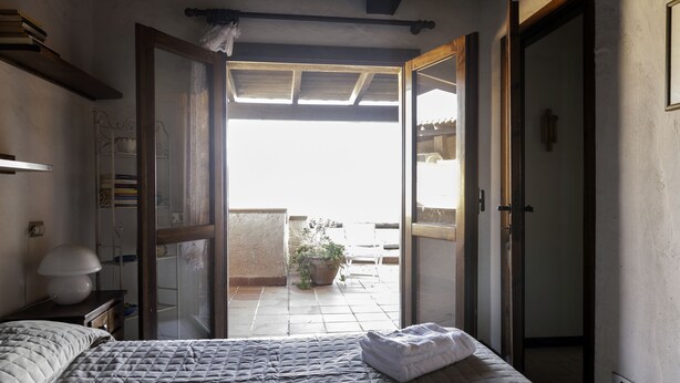 A bedroom with a bed, bedside table, lamp, and a view of a balcony with a potted plant. - Aria di Vacanza - Corallina (Loiri Porto San Paolo)