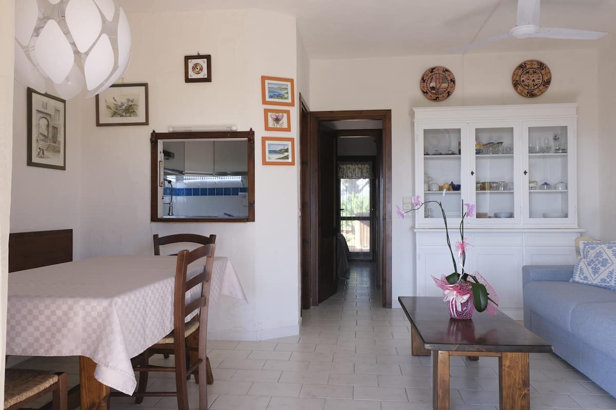 Ein Essbereich mit Tisch und Stühlen, eine durch ein Fenster sichtbare Küche, ein Wohnzimmer mit Sofa und Schrank, sowie ein Flur, der in ein weiteres Zimmer führt.